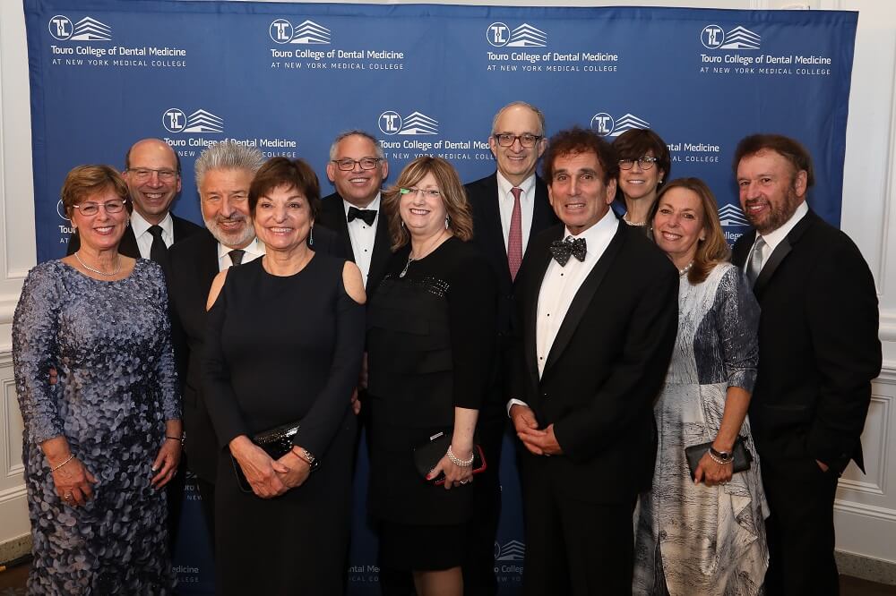 Men and women dressed in formal wear standing in front of a Touro College of Dental Medicine banner.