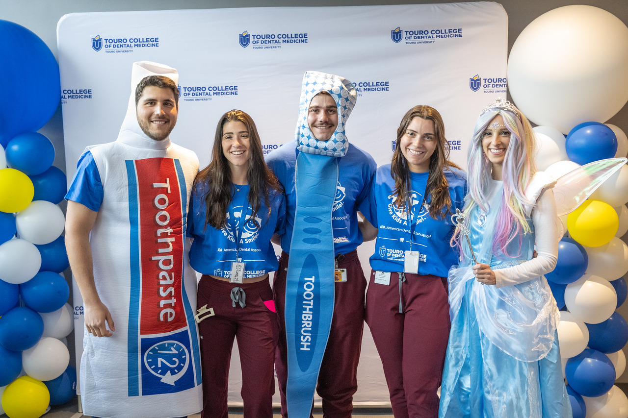 Five dental students standing together at the 2023 Give Kids a Smile program at Touro College of Dental Medicine. They are in front of a TCDM step-and-repeat with blue yellow and white balloon pillars on either side. Three of the students are in dentistry-related costumes - a tube of toothpaste, a toothbrush and the tooth fairy.