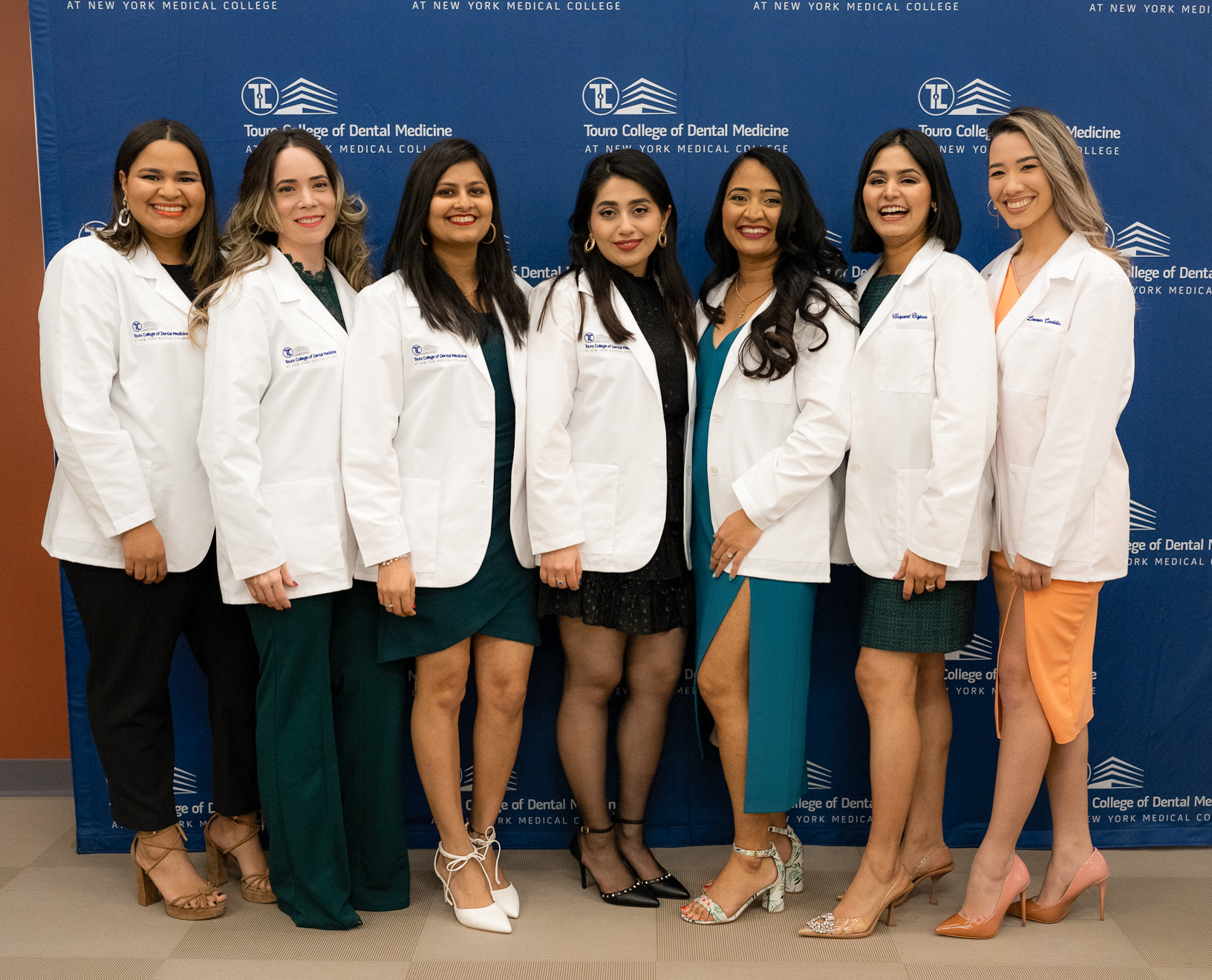 Seven women, the first cohort, in the advanced standing international dentist program stand in their white coats in front of a Touro College of Dental Medicine step-and-repeat.