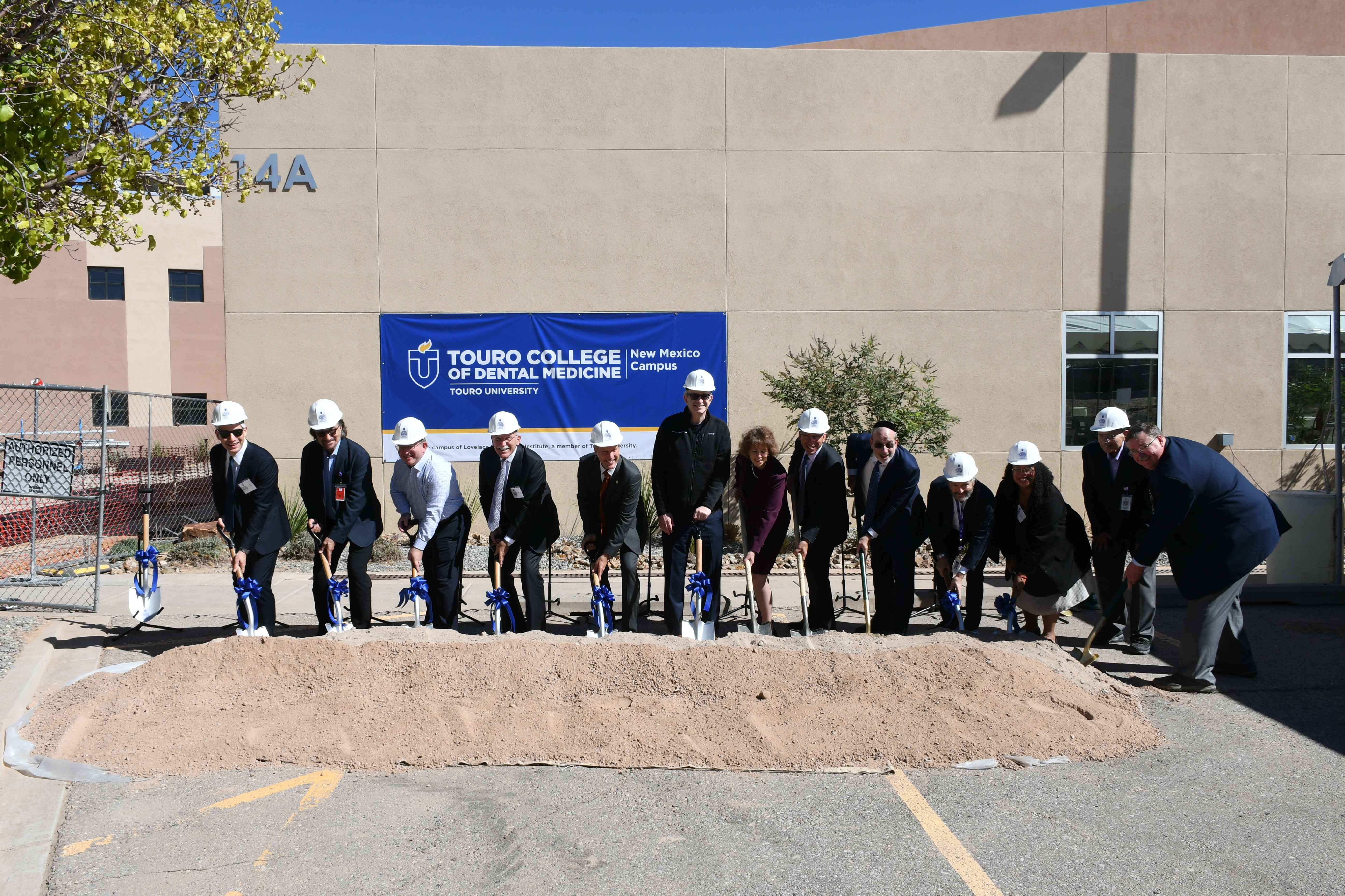 Touro leadership in suits and hard hats, holding shovels in dirt in lot, at the location of where the new Touro College of Dental Medicine front of where the future undergraduate dental clinical training facility will be built.