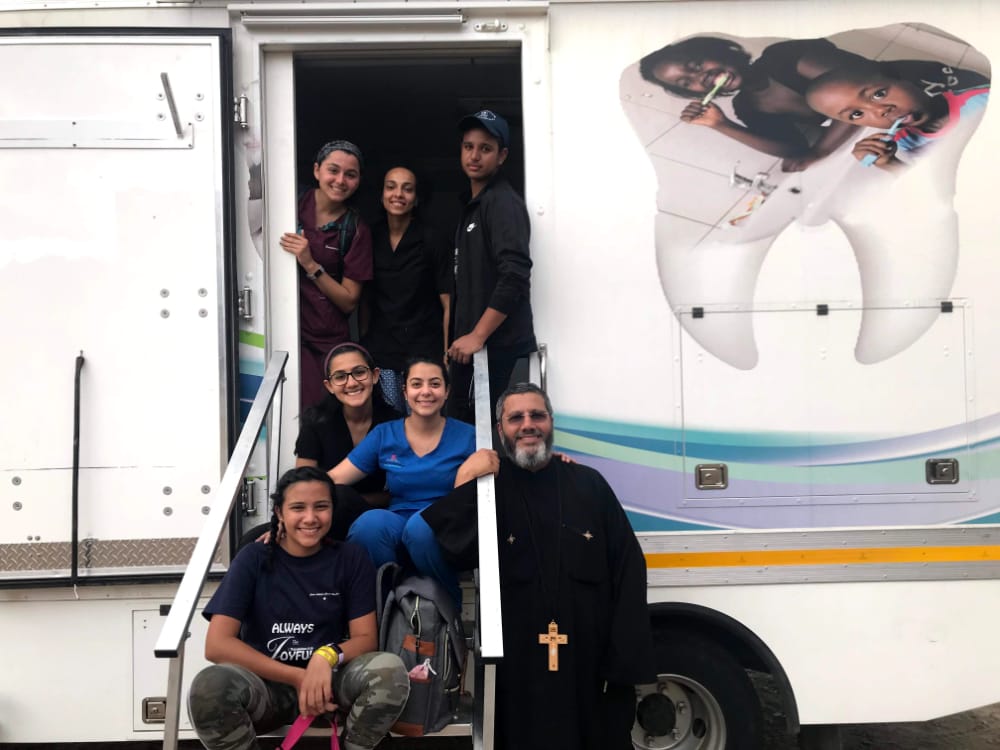 Simona Girgis posing with other TCDM student volunteers and a priest, outside of a volunteer trailer