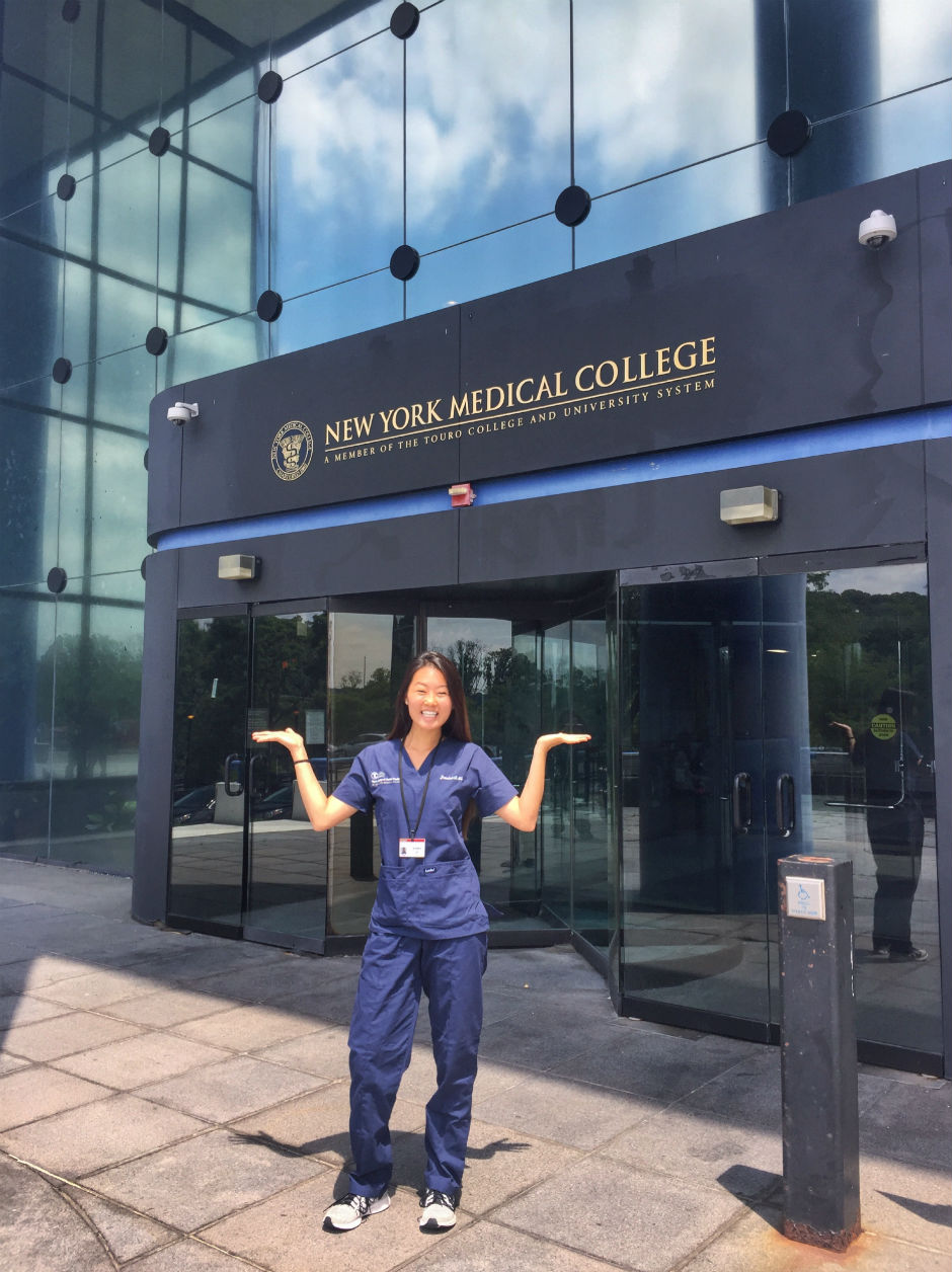 Jessica Li in navy scrubs stands in front of the TCDM entrance with the NYMC signage above the door. 